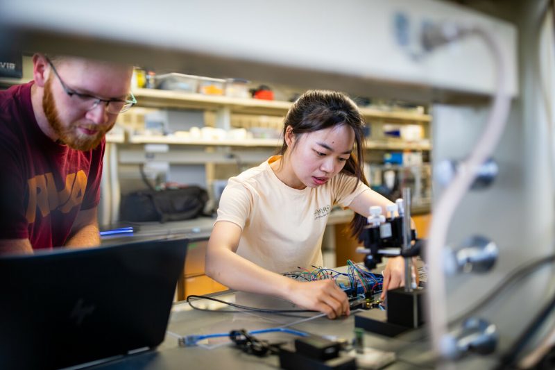 Two researchers in a lab, one looking at a computer, the other testing a connection to a board.