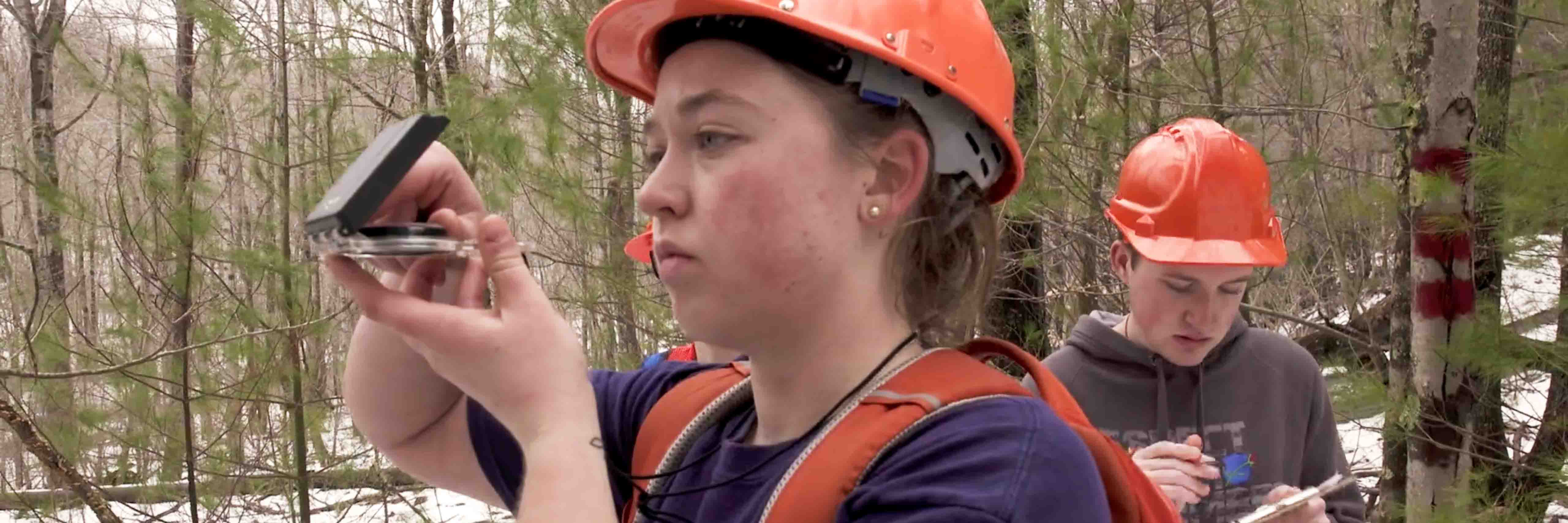 Two students in a light snow covered forest wearing hard hats, one holds a compass up to eye level while the other writes on a clipboard