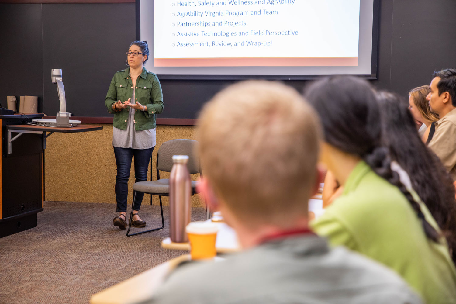 A woman stands at the front of a room in front of a slideshow presentation and talks.