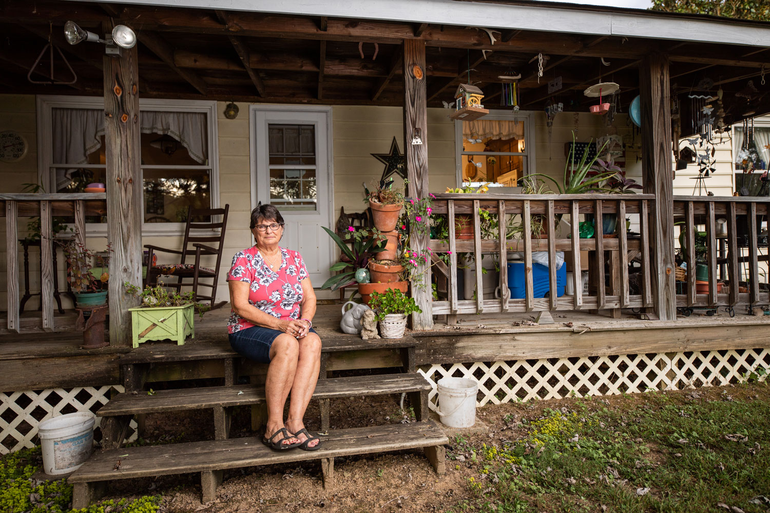 A woman sits on a porch filled with plans and wind chimes outside a house.