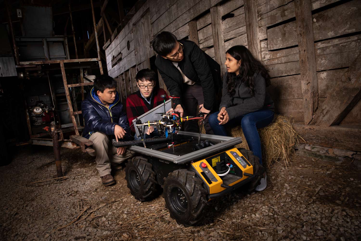 Four students crowd around a drone and ground vehicle while sitting inside a barn.