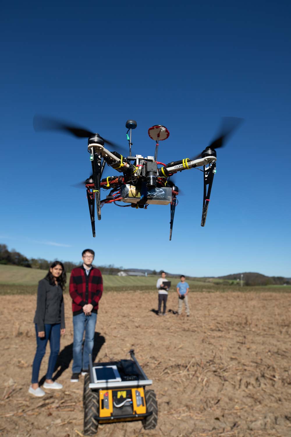 A drone hovers in the center of the phone. In the background students stand near a ground vehicle while the drone operators stand further back.