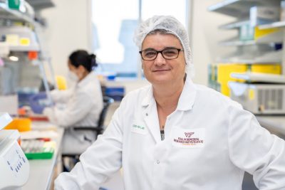 A woman in a white lab coat and with a hair net stands in her medical laboratory.