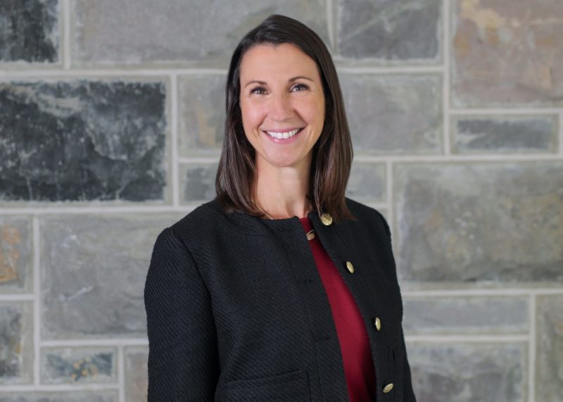 A person with shoulder-length dark hair stands smiling before a wall of grey stone.
