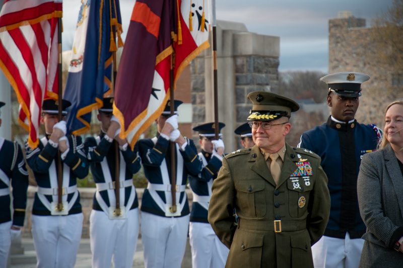 Maj. Gen. William Seely (at enter) smiles while standing in his Marine uniform in front of a group of cadets carrying flags.