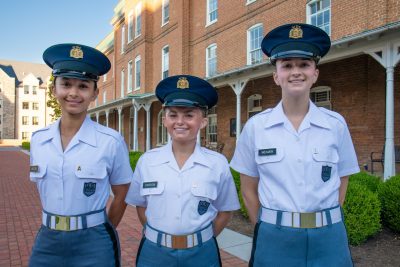 Three women cadets stand in uniform smiling in front of the brick building Lane Hall on a brick sidewalk. 