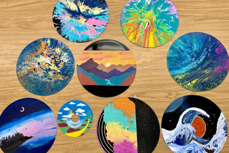 Several painted vinyl records are displayed on a wooden table, each decorated by student artists. The designs include vibrant abstract paint pours, a mountain landscape at sunset, a night shoreline scene, a painted 45 record with a "Wizard of Oz" inspired theme, and a multicolored split abstract piece.