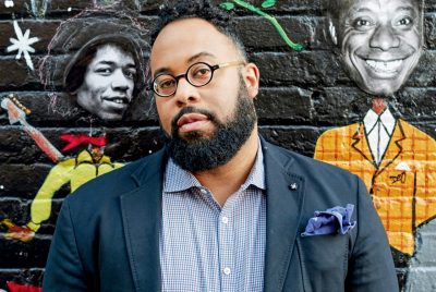 Kevin Young is a black man with a beard and glasses, wearing a navy blazer and checkered button-up shirt, standing in front of a brick wall painted black with images of Black artists.