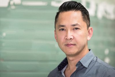 Author Viet Thanh Nguyen is a Vietnamese-American man wearing a patterned, collared, buttoned-up shirt standing in front of a pale green brick wall.