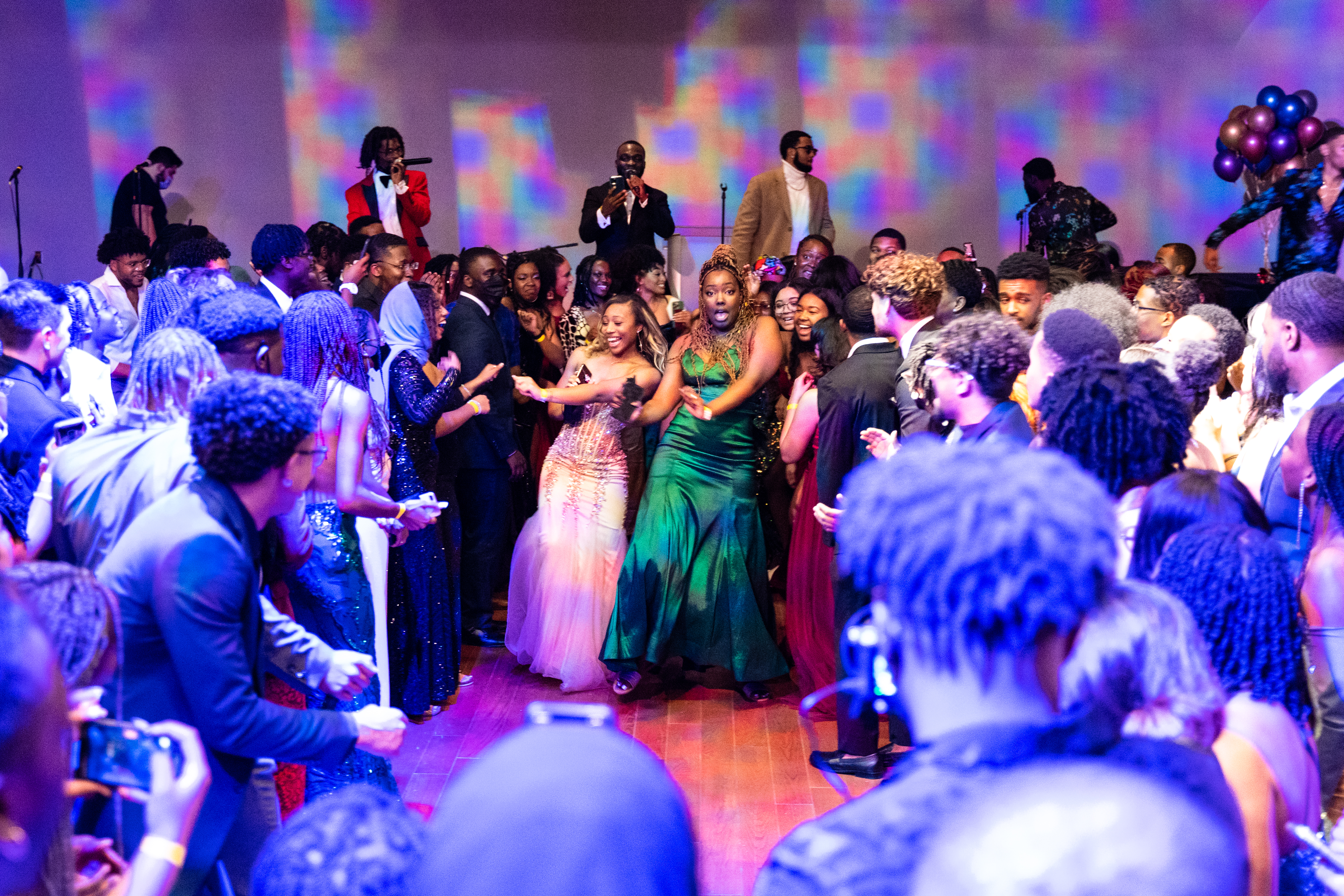 Students gather in celebration at the Black Excellence Gala. Two female students are centered, smiling and dancing in a large group of  students. All are in formal wear. 